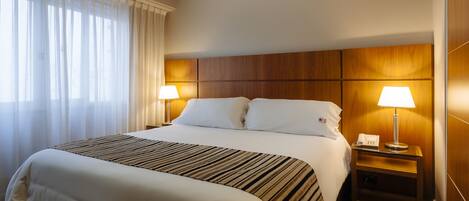 A hotel room with a large bed, two bedside lamps, a wooden headboard, and a window with sheer curtains.