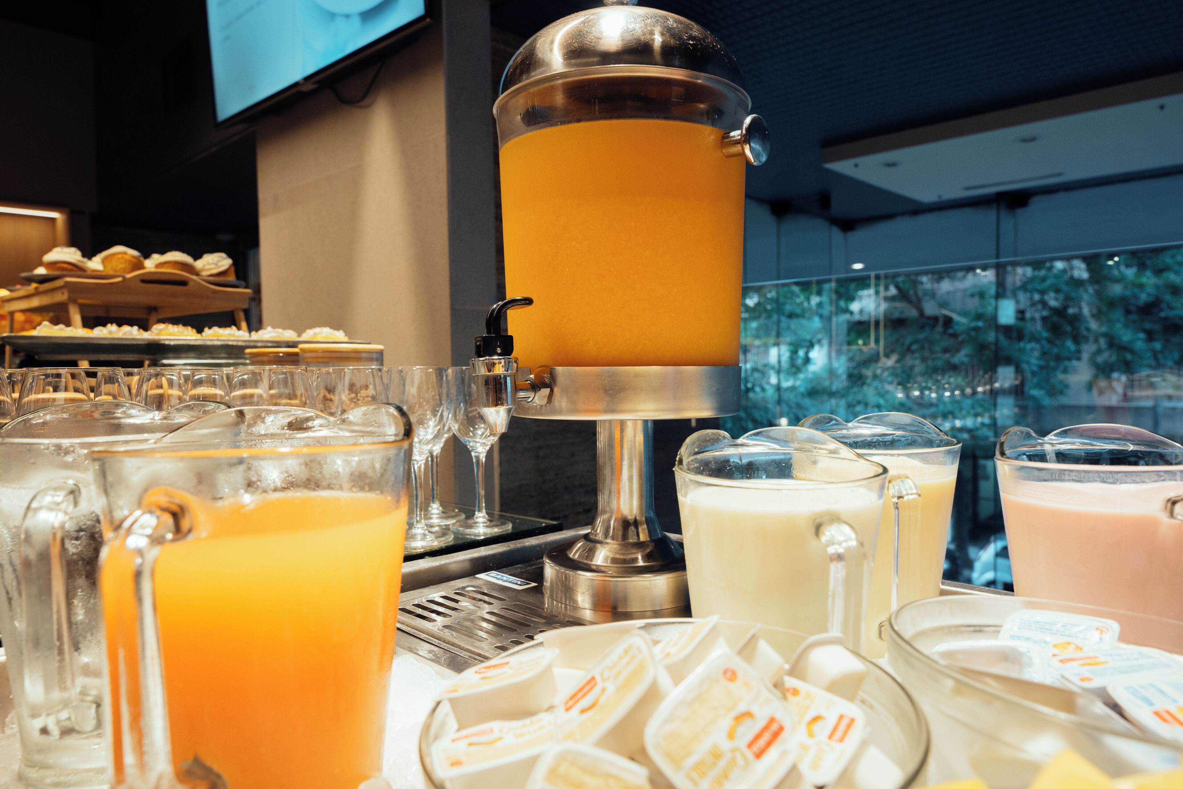 A beverage station with multiple dispensers and glasses, and a buffet area with pastries in the background.