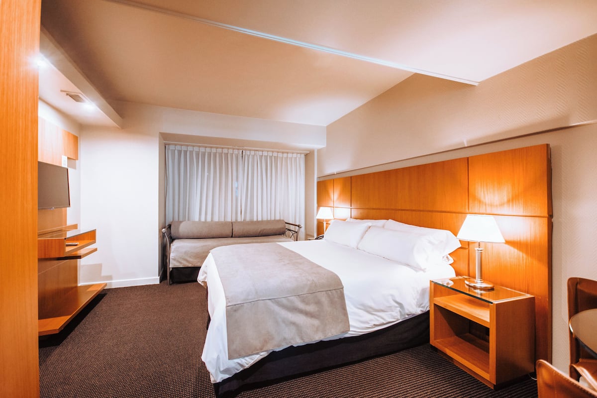 A hotel room with a bed, bedside table, lamp, and a view of the outside through a window.