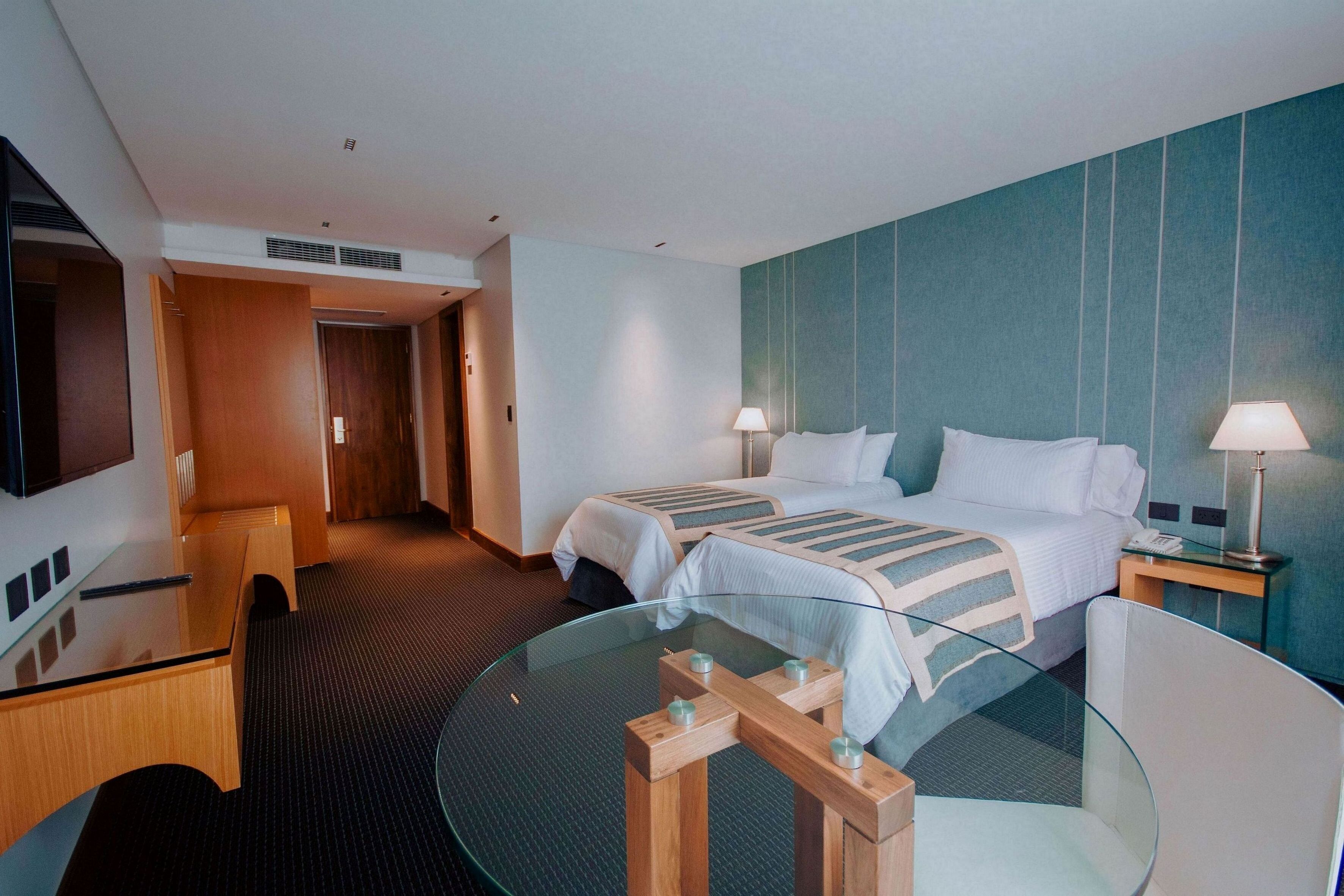 Habitación de hotel con dos camas, una mesa de vidrio, un escritorio y un televisor.