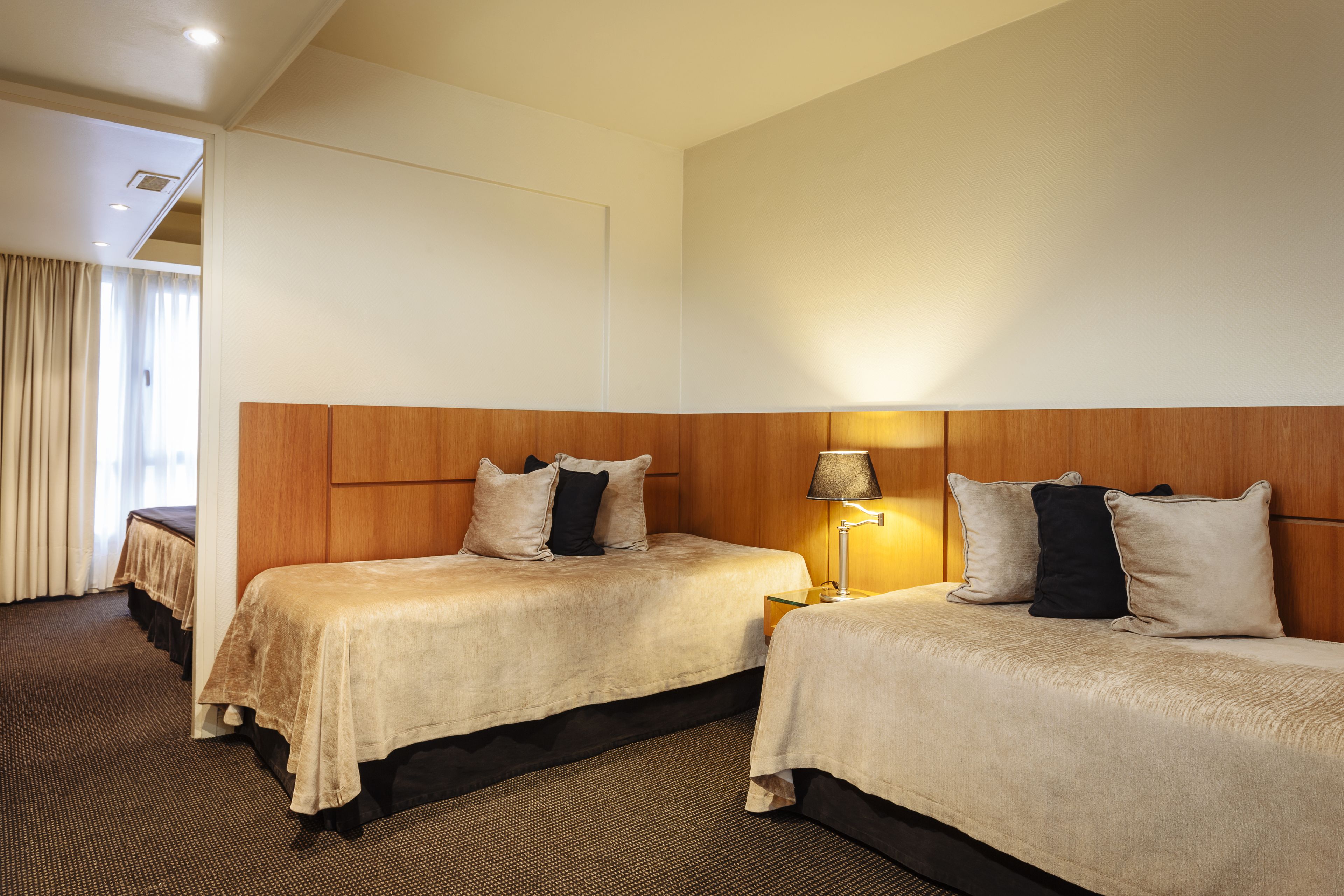 A hotel room with two beds, a wooden headboard, a lamp, and a view of the outside through a window with curtains.
