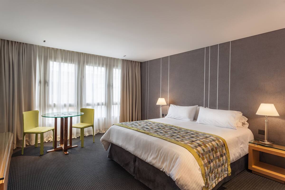 A hotel room with a large bed, two chairs, a small table, and a nightstand with a lamp.