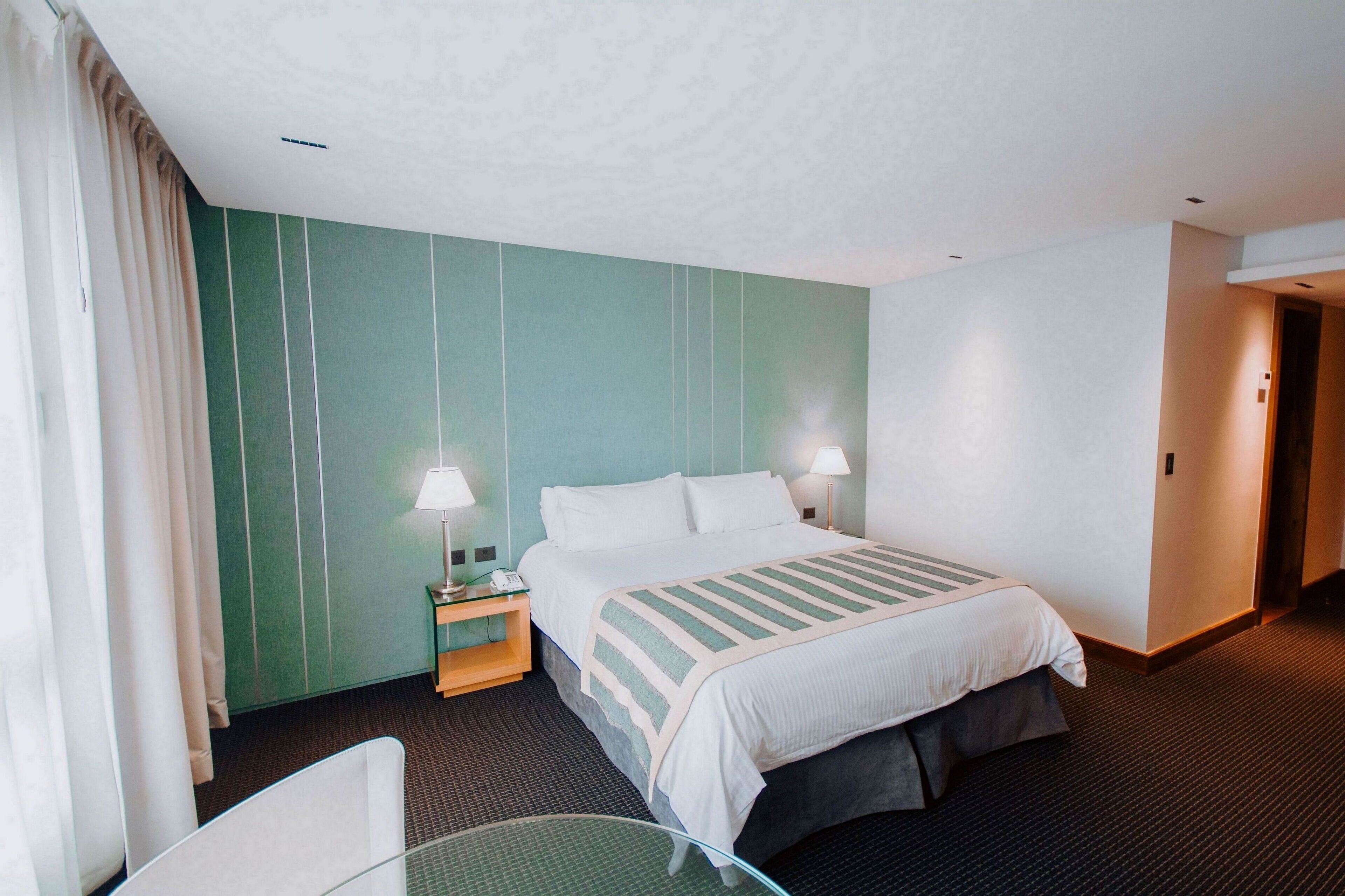 A hotel room with a bed, two bedside lamps, a nightstand, a glass table, and a curtain.