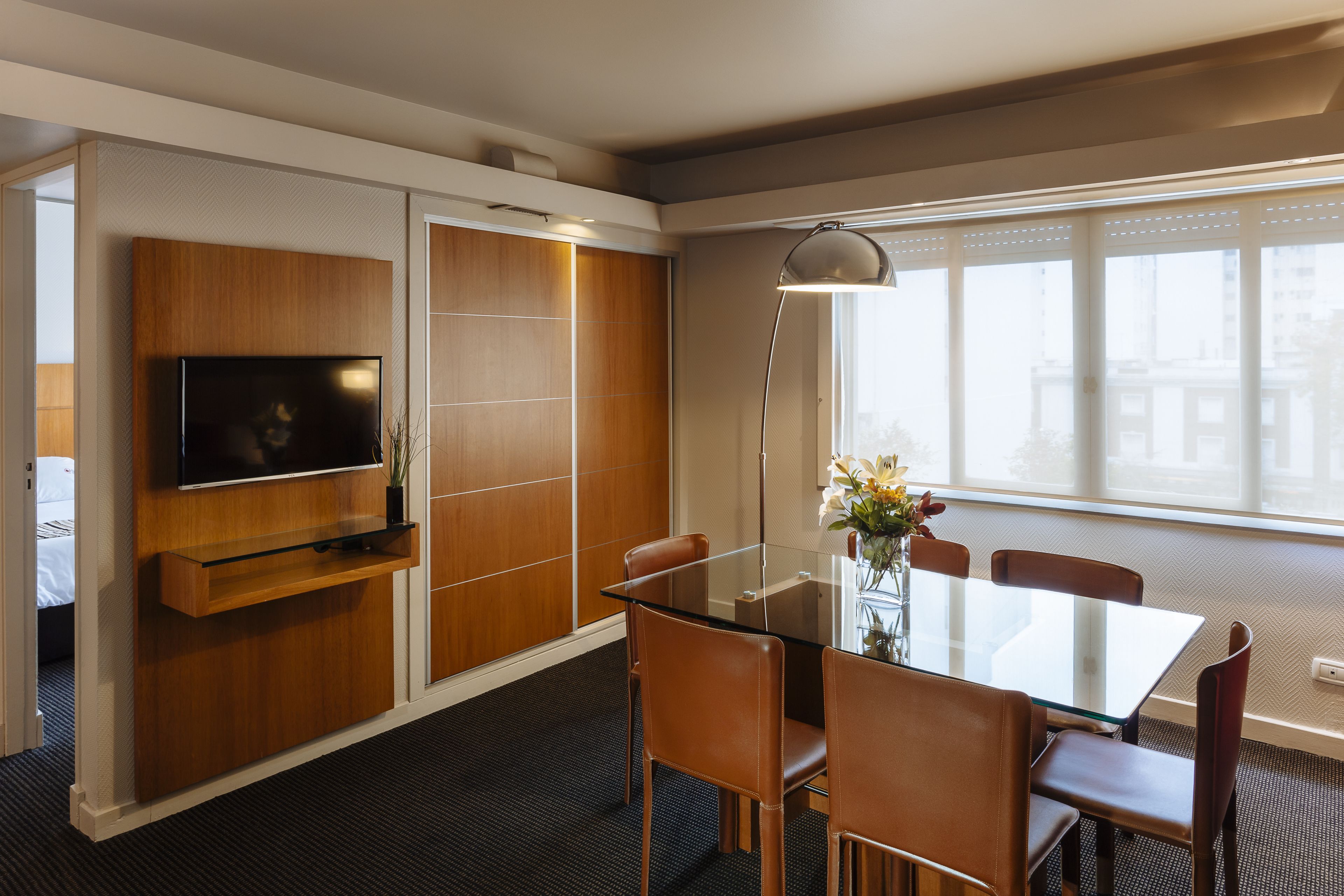 A modern hotel room with a glass dining table, brown leather chairs, a flat-screen TV mounted on a wooden panel, and a large window offering a city view.