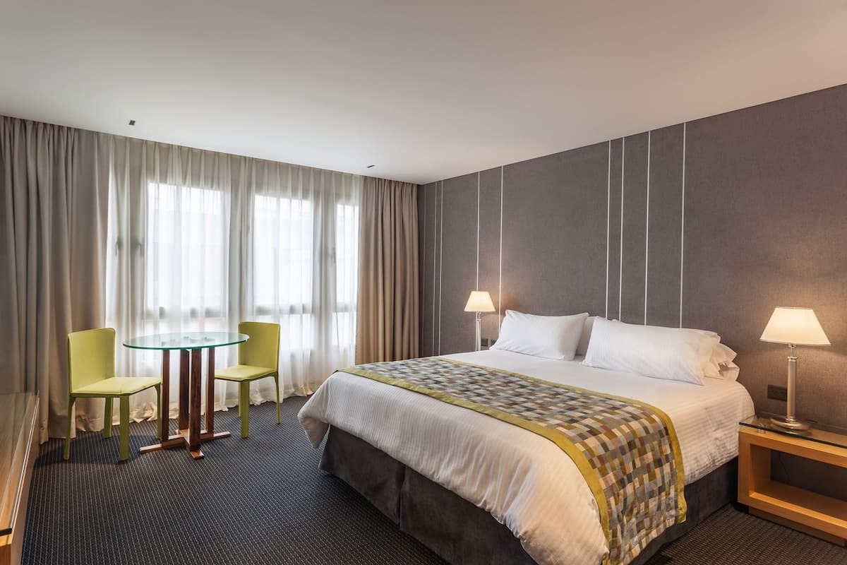 A hotel room with a bed, two bedside lamps, a small table with two chairs, and a large window with curtains.