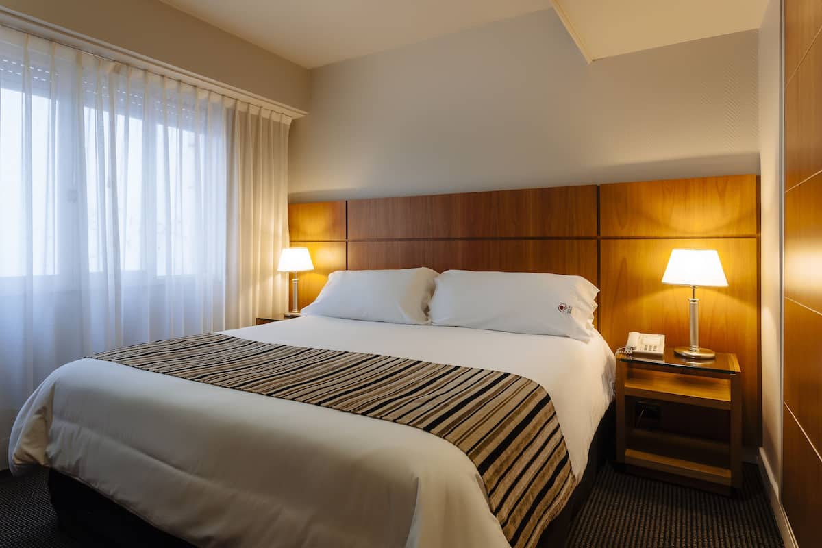 A hotel room with a large bed, two bedside lamps, a wooden headboard, and a window with sheer curtains.