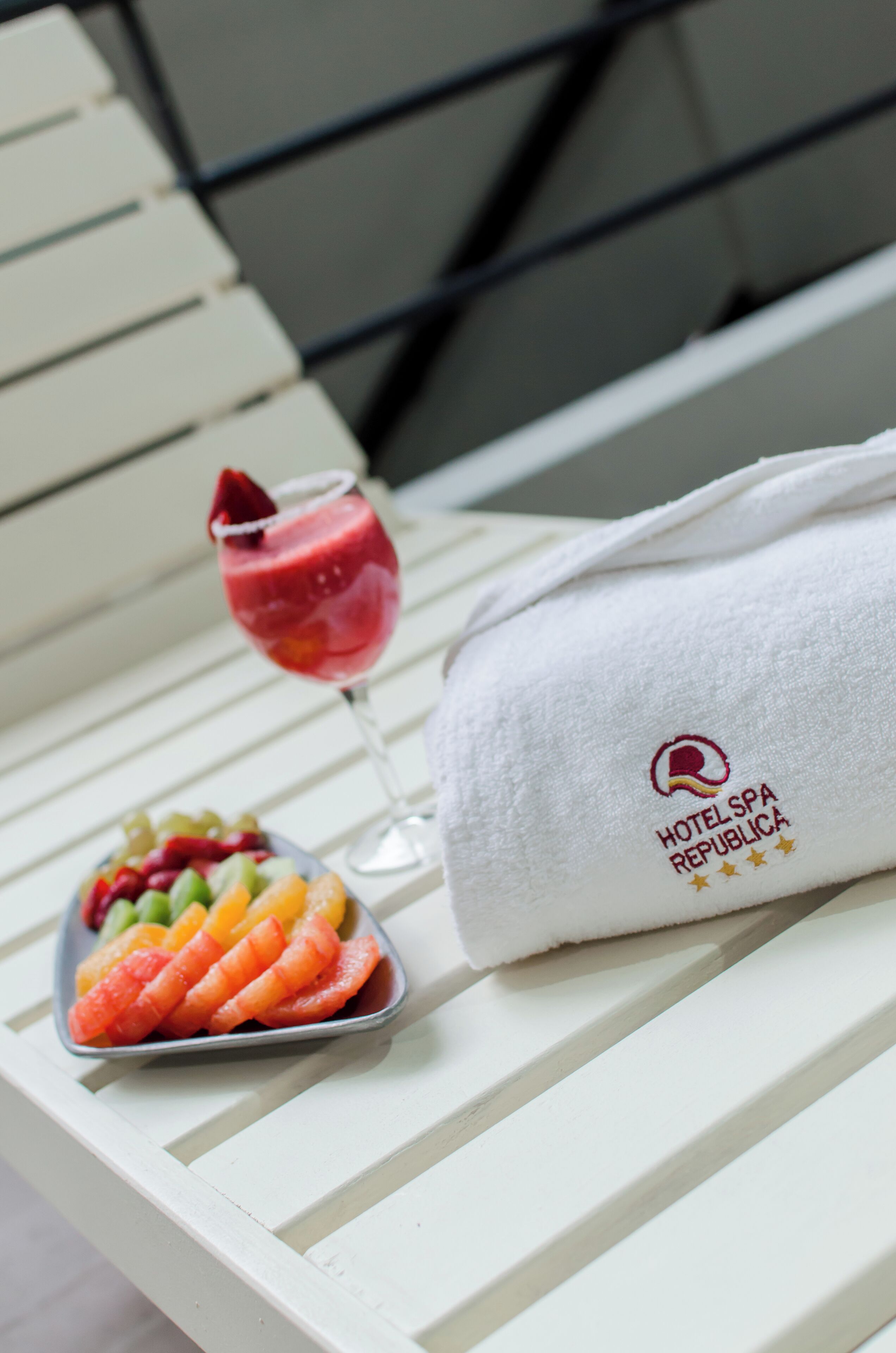 A white towel with a logo and text, a glass of red drink, and a plate of fruit on a white chair.