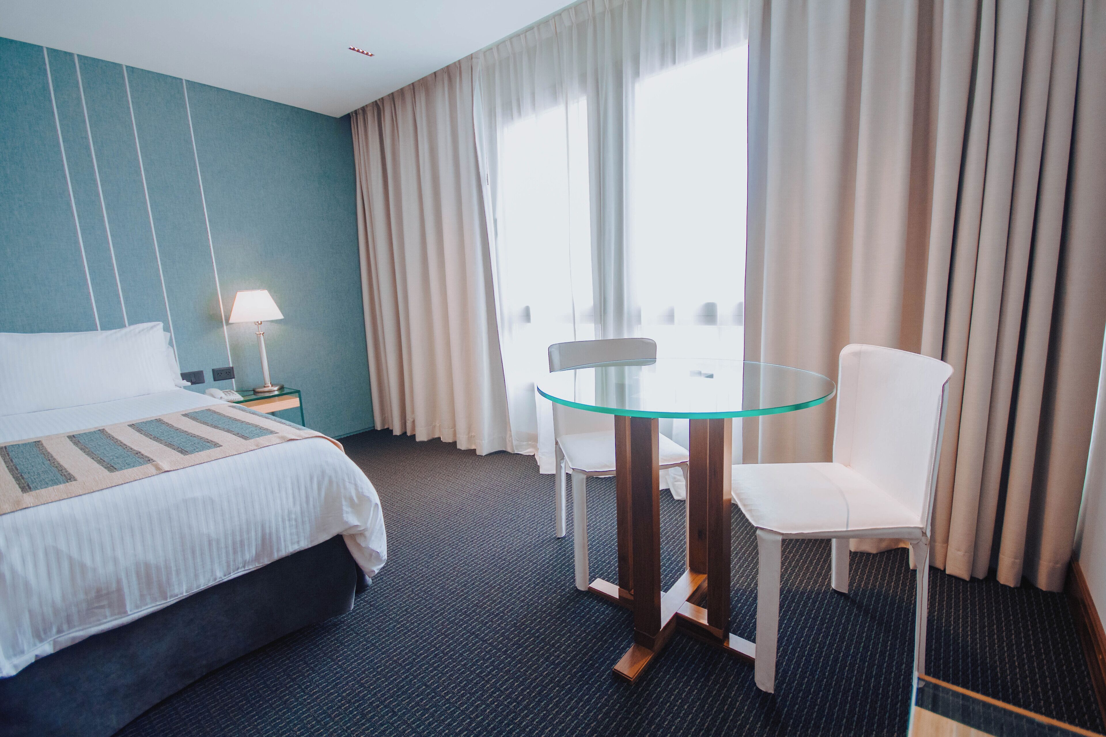 Habitación de hotel con una cama, una mesa de vidrio, dos sillas blancas, una lámpara y vistas al exterior a través de amplios ventanales.