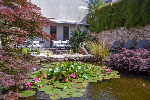A serene garden with a pond, lily pads, and pink water lilies. There are lounge chairs and a seating area with a stone wall and greenery.