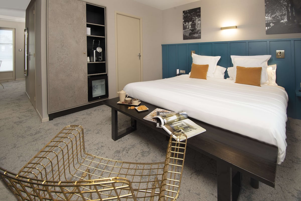 A hotel room with a large bed, a bedside table, a gold wire chair, a wardrobe, and a desk with a magazine.