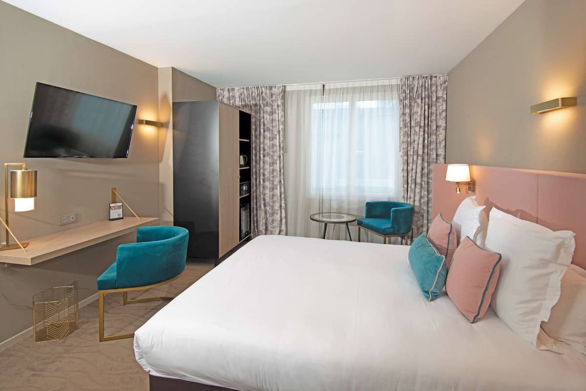 A hotel room with a large bed, a TV mounted on the wall, a small table, two chairs, and a bookshelf.