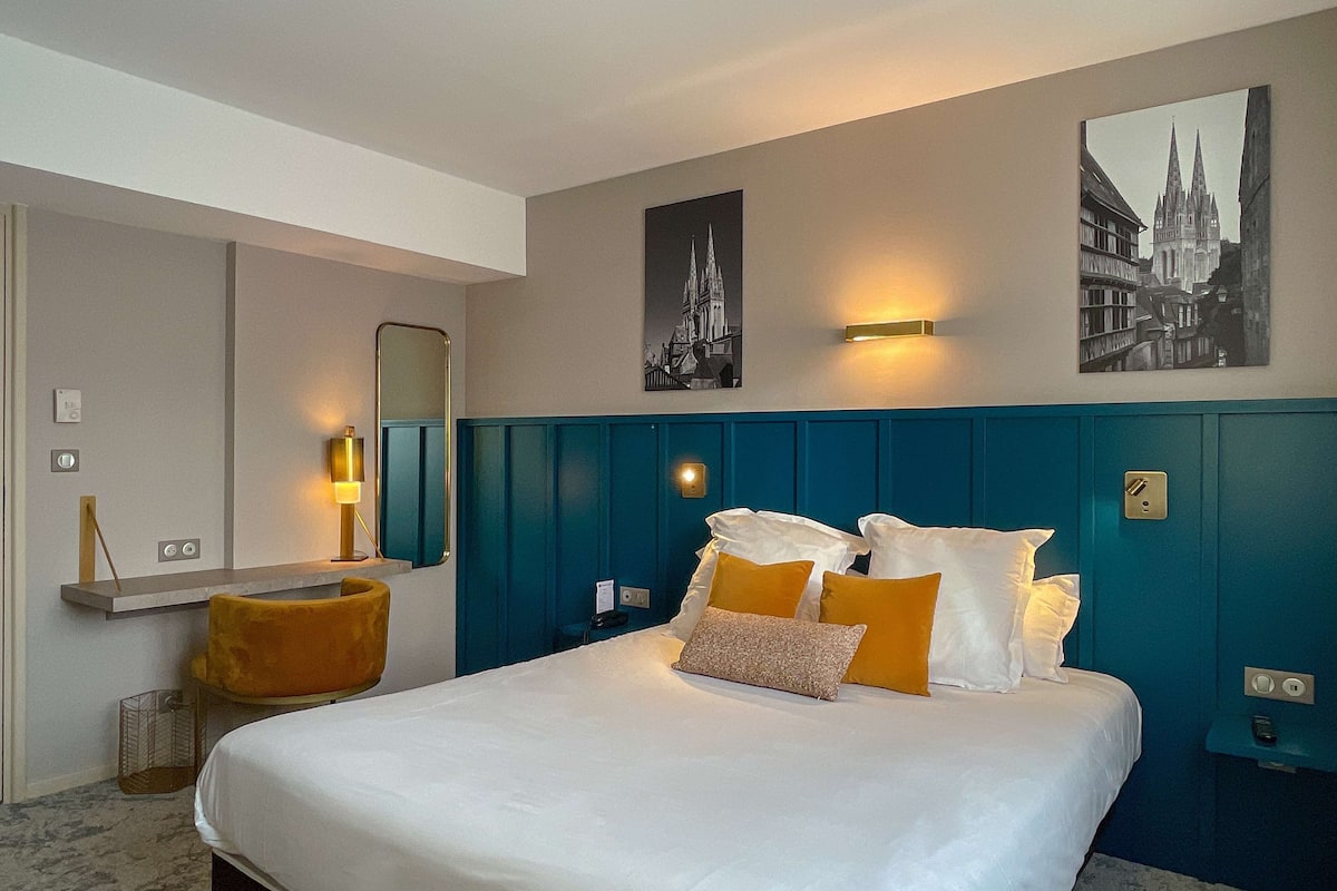 A hotel room with a large bed, a wooden chair, a mirror, and two wall-mounted light fixtures.