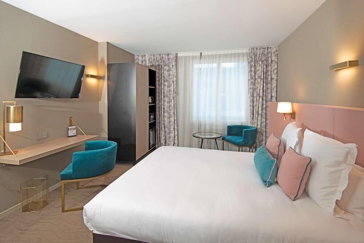 A hotel room with a large bed, a TV mounted on the wall, a desk with a chair, and a small table with chairs.