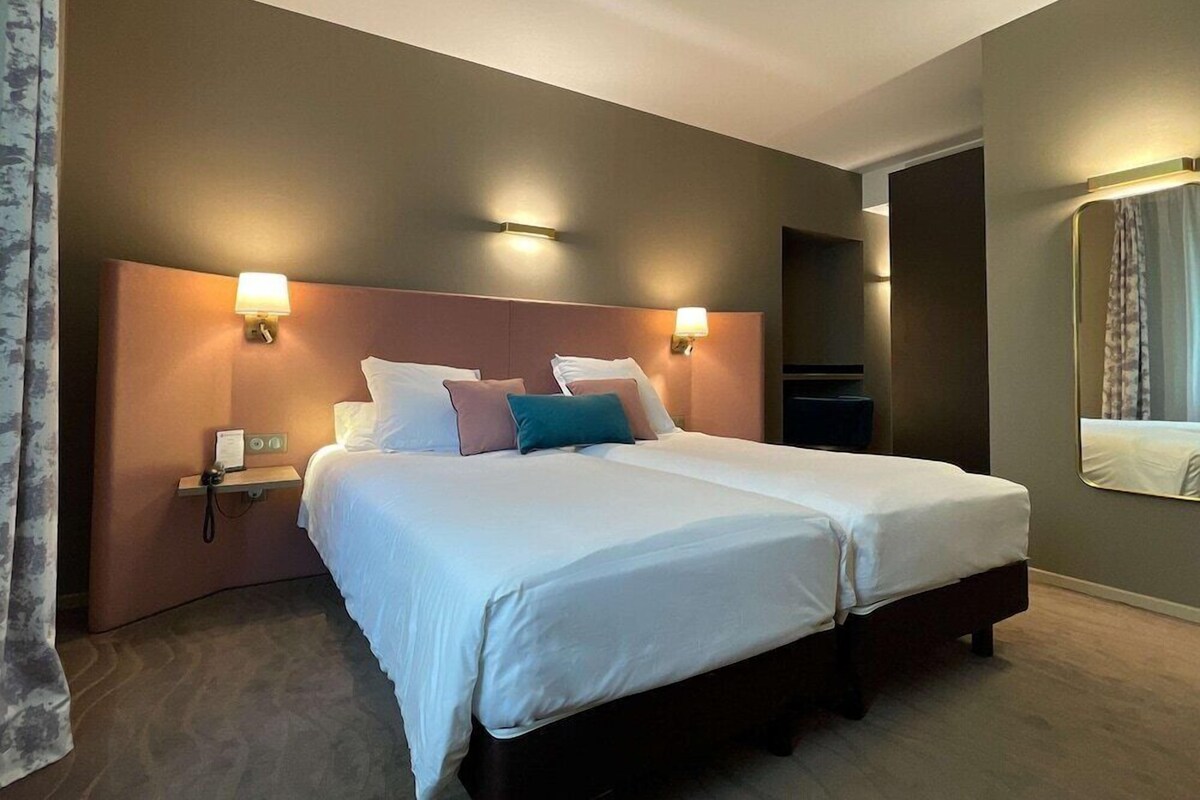 A double bed with white linens and multiple pillows, a bedside table with a lamp, and a wall-mounted TV.