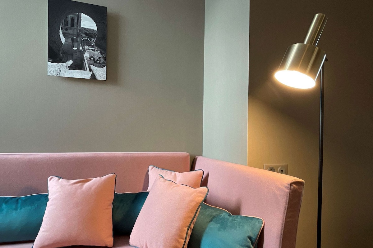 A pink sofa with teal cushions, a black and white framed picture on the wall, and a floor lamp with a metallic shade.