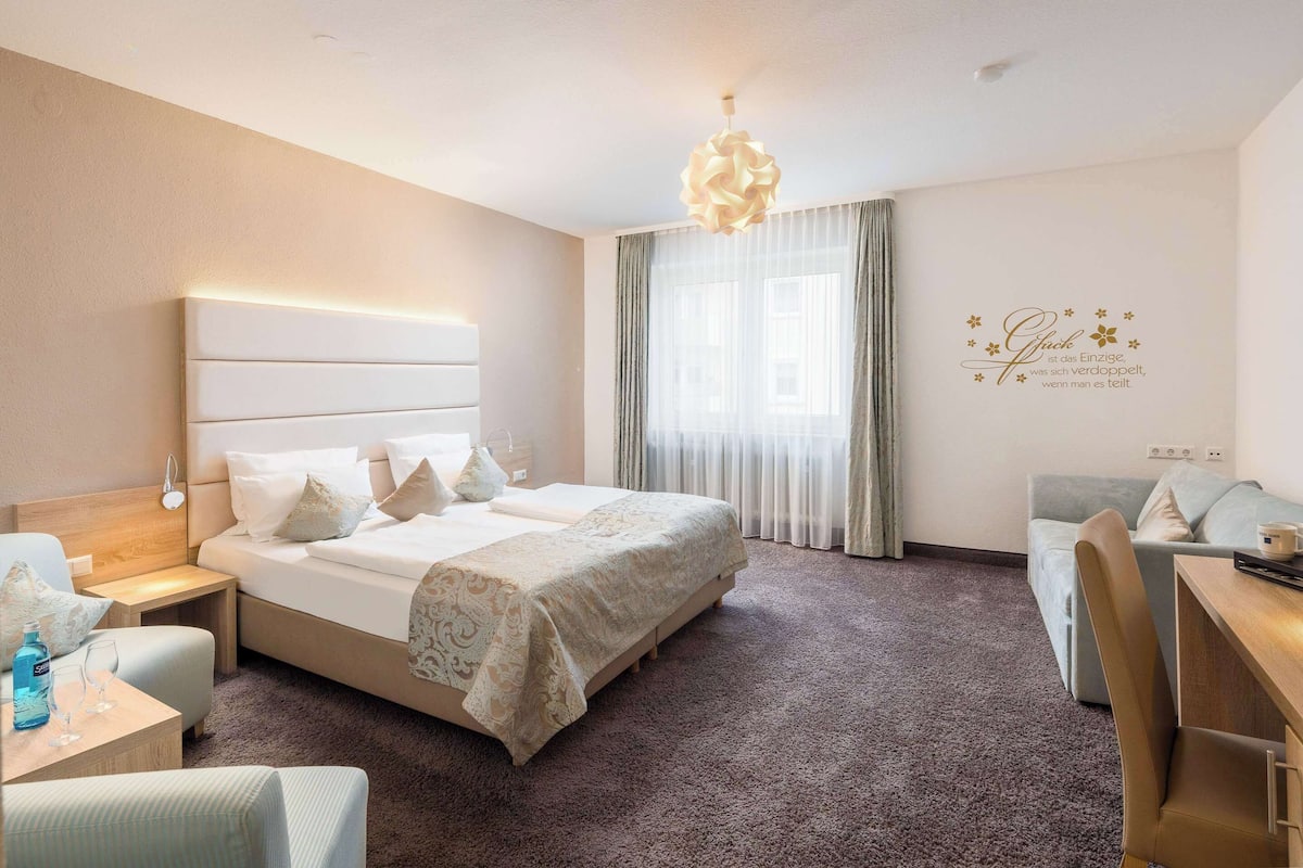 A hotel room with a large bed, bedside tables, a desk, a chair, and a sofa.