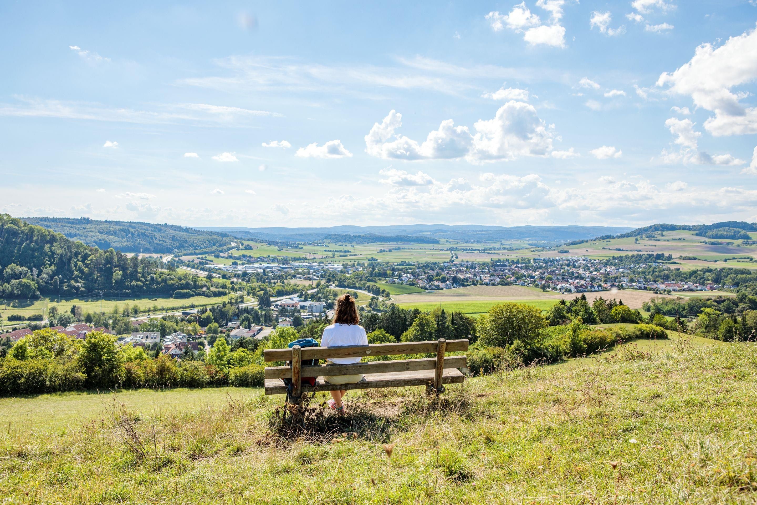 Une personne assise sur un banc, d’où l’on a une vue sur une ville et un paysage vallonné.