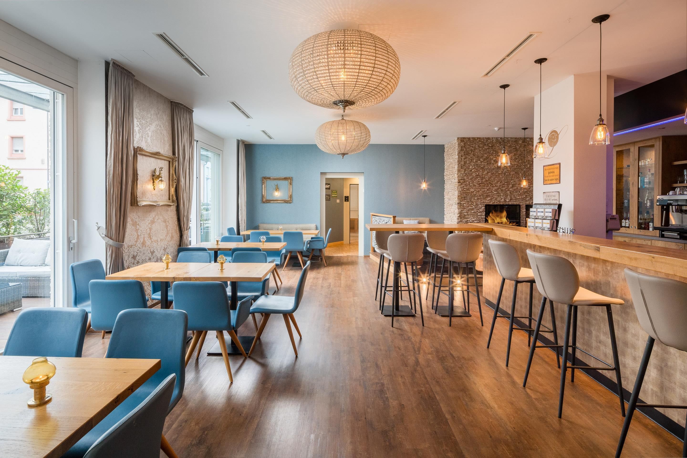 Un interior de restaurante moderno con mesas de madera y sillas azules, una zona de bar con taburetes y una pared de piedra.