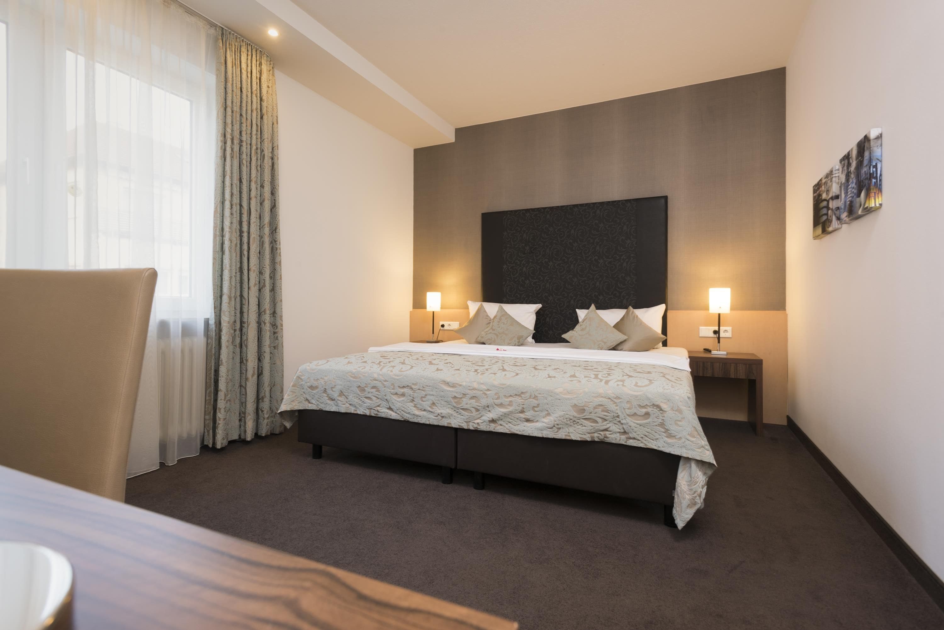 A hotel room with a large bed, two bedside tables with lamps, a chair, and a view of the outdoors through the window.