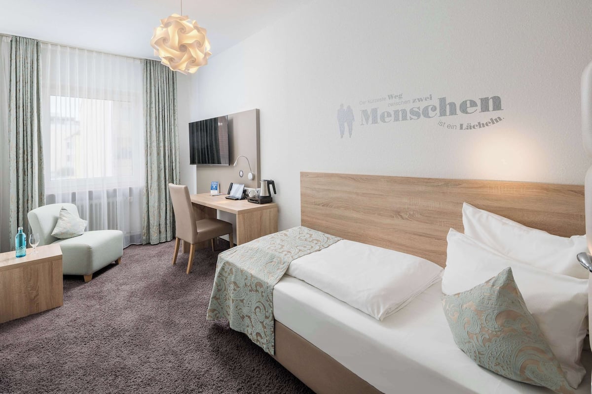 A hotel room with a bed, a desk, a chair, a TV, and a wall with a motivational quote.