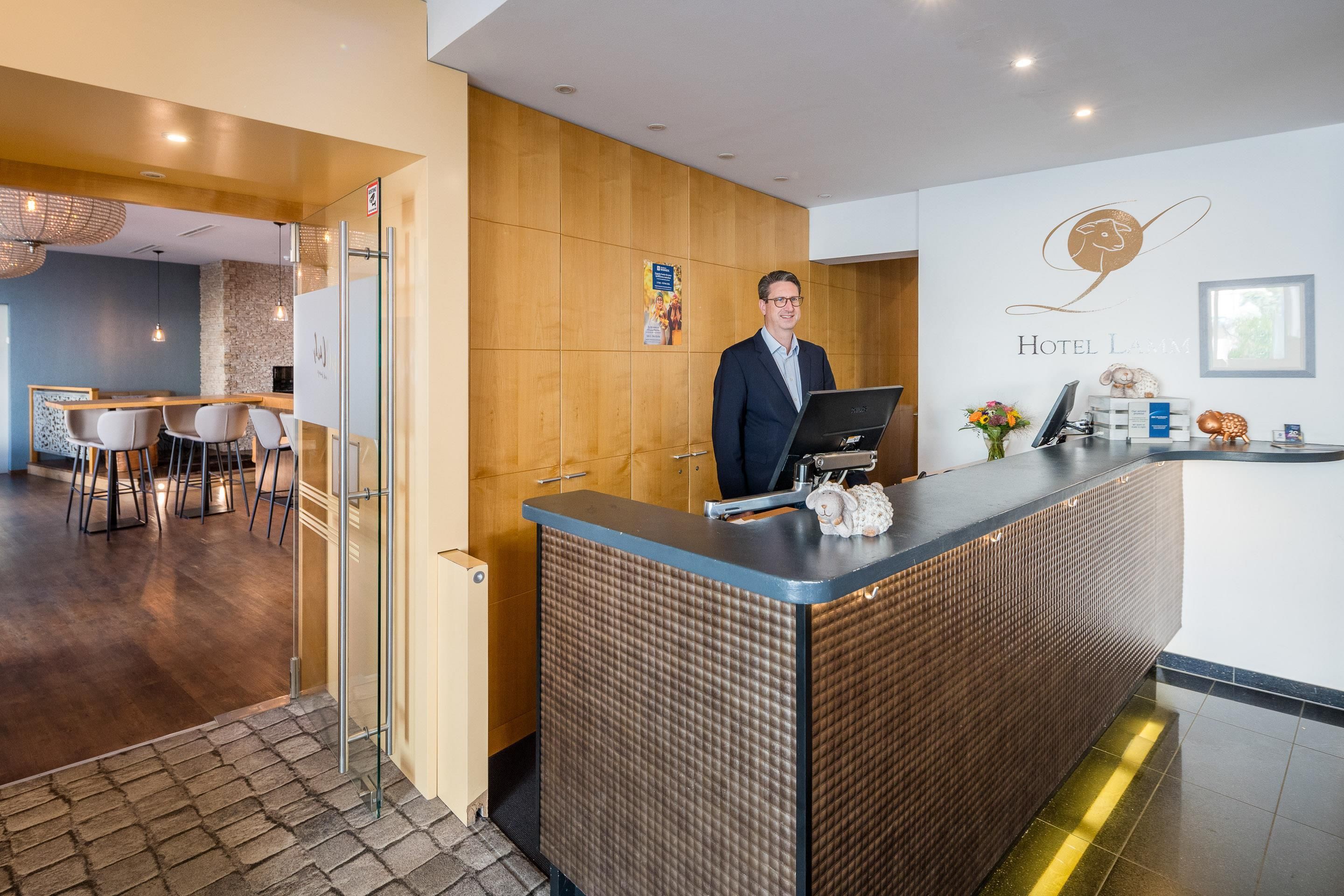 Un espace d’accueil d’hôtel avec un comptoir en bois, un homme derrière le comptoir, et un bar avec des tabourets.