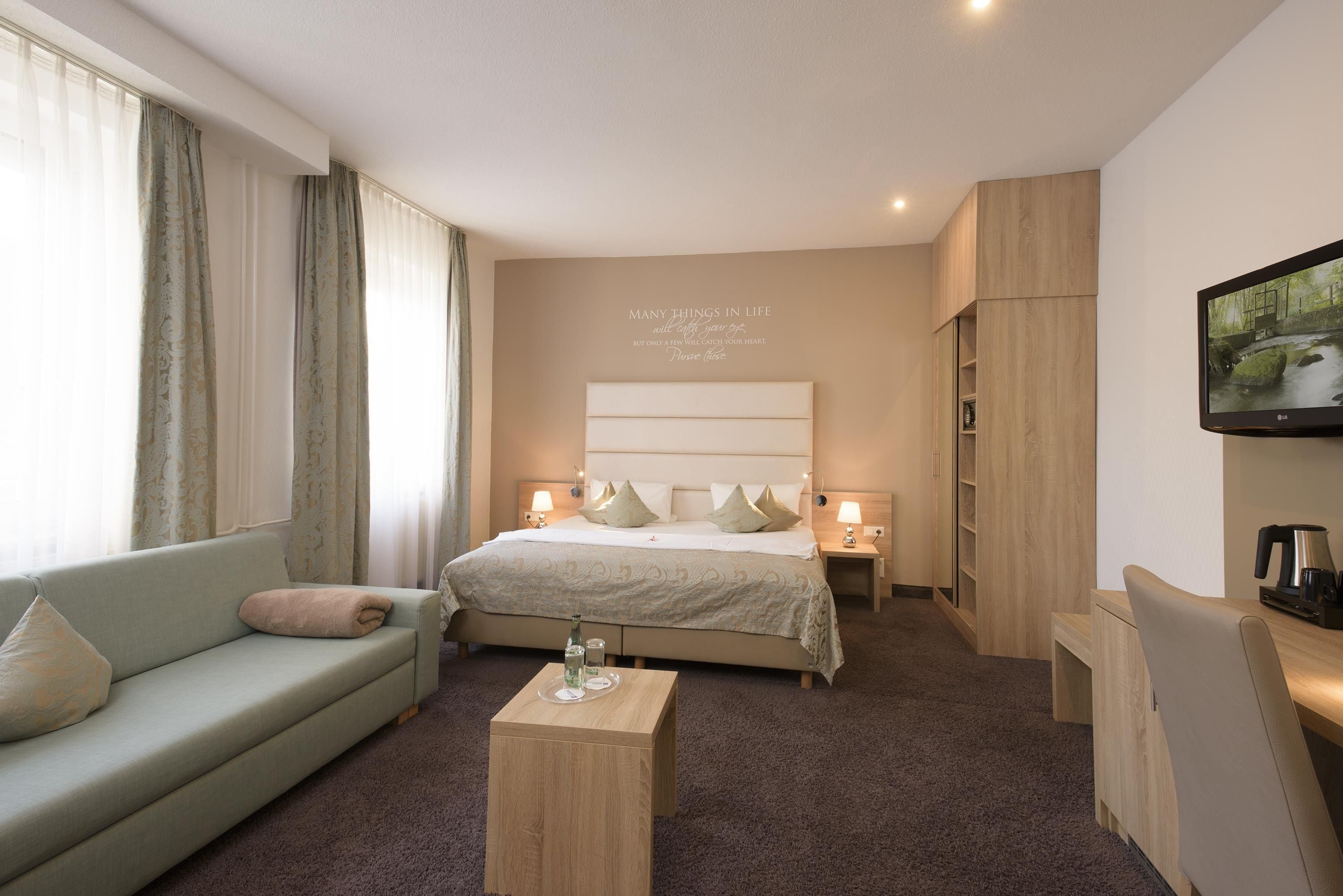 Una camera d'albergo con un letto, un divano, un tavolino da caffè, una televisione e una parete con una citazione motivazionale.