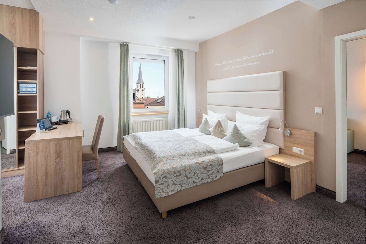 A hotel room with a bed, bedside table, desk, and a view of a church through a window.