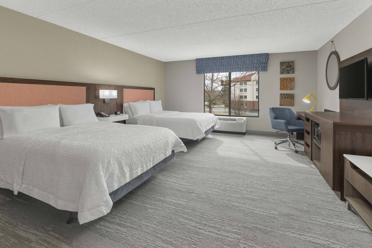 Une chambre d’hôtel avec deux lits, un bureau, une chaise, une télévision et une fenêtre donnant sur des immeubles.