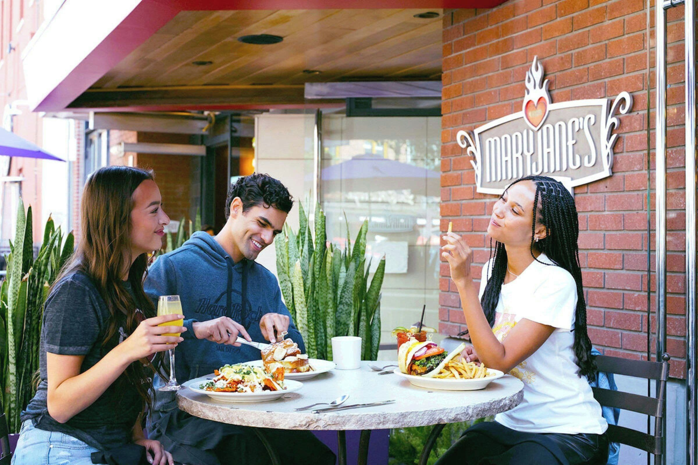 Three people dining at an outdoor table with a brick wall and a sign that reads 'Mary Jane's'.