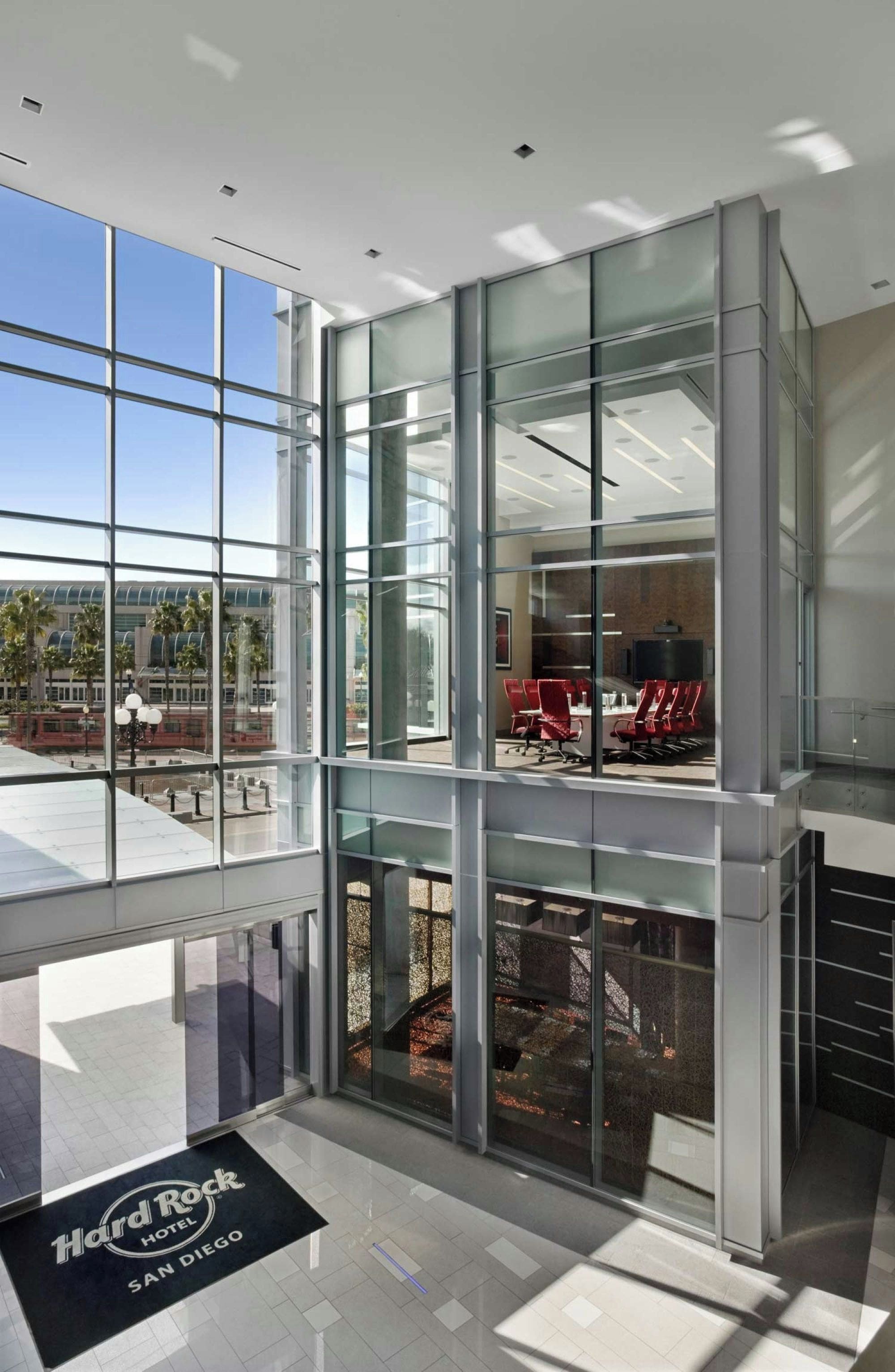 A modern hotel lobby with large glass windows, a Hard Rock Hotel sign, and a view of the street outside.