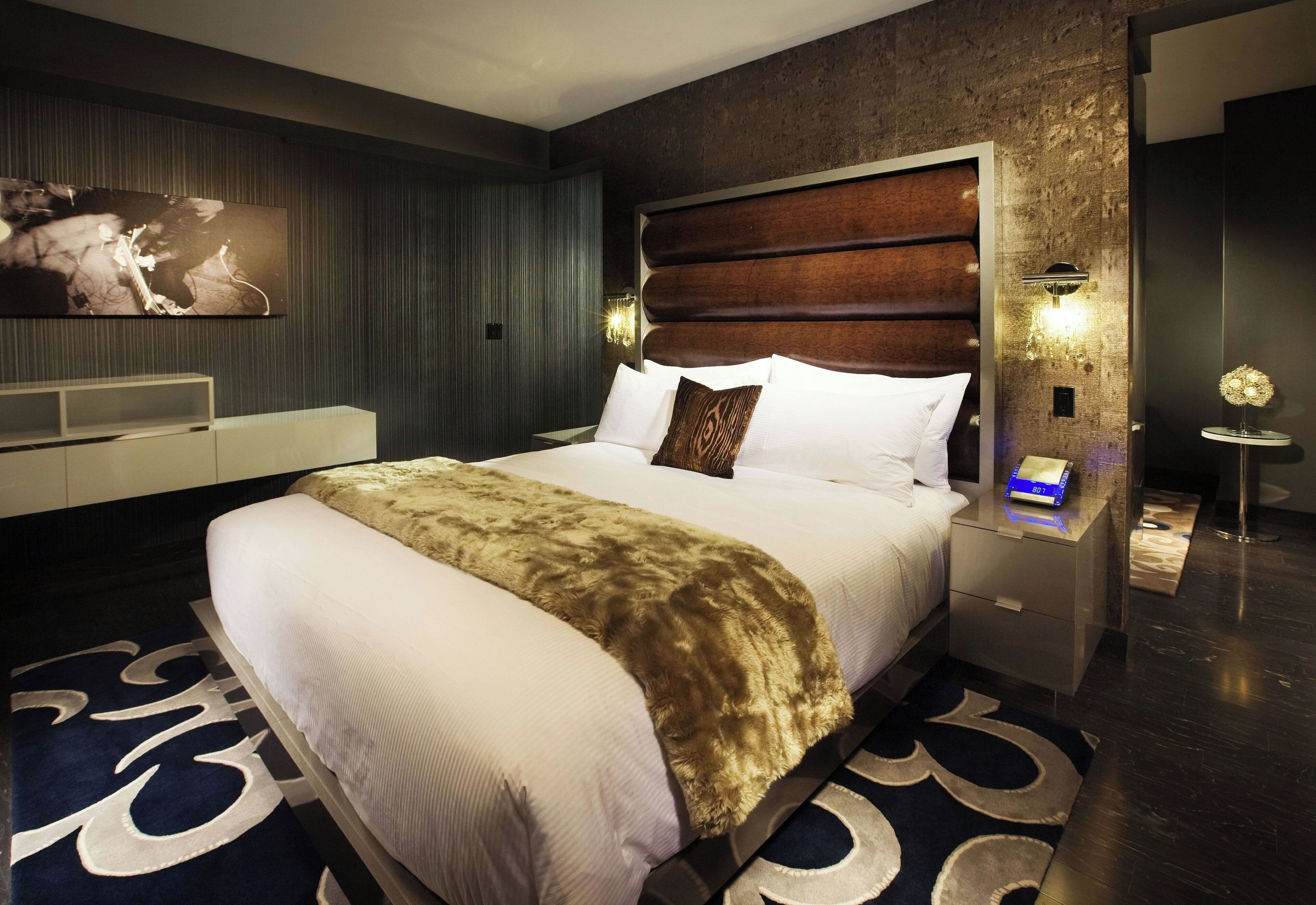 A modern hotel room with a large bed, a flat-screen TV mounted on the wall, a nightstand with a lamp, and a decorative rug.