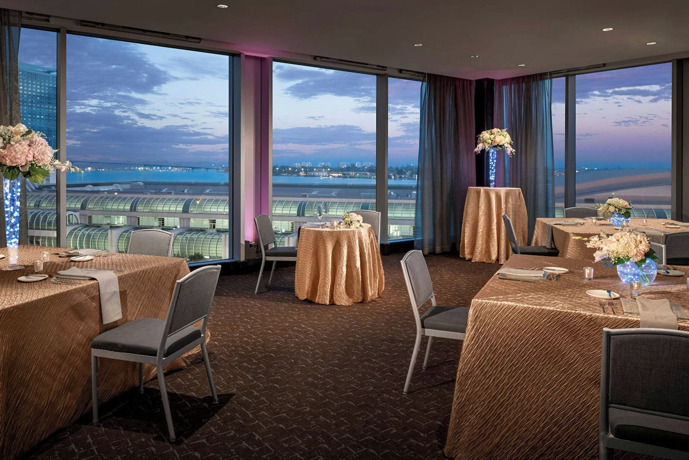 Un comedor de hotel con mesas dispuestas para el servicio, grandes ventanales con vistas a un paisaje urbano y al agua, y suelo de ladrillo.