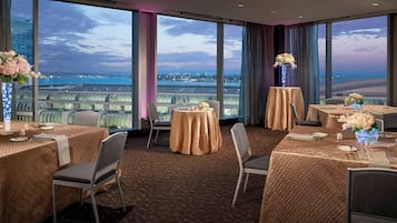 Un comedor de hotel con mesas dispuestas para el servicio, grandes ventanales con vistas a un paisaje urbano y al agua, y suelo de ladrillo.