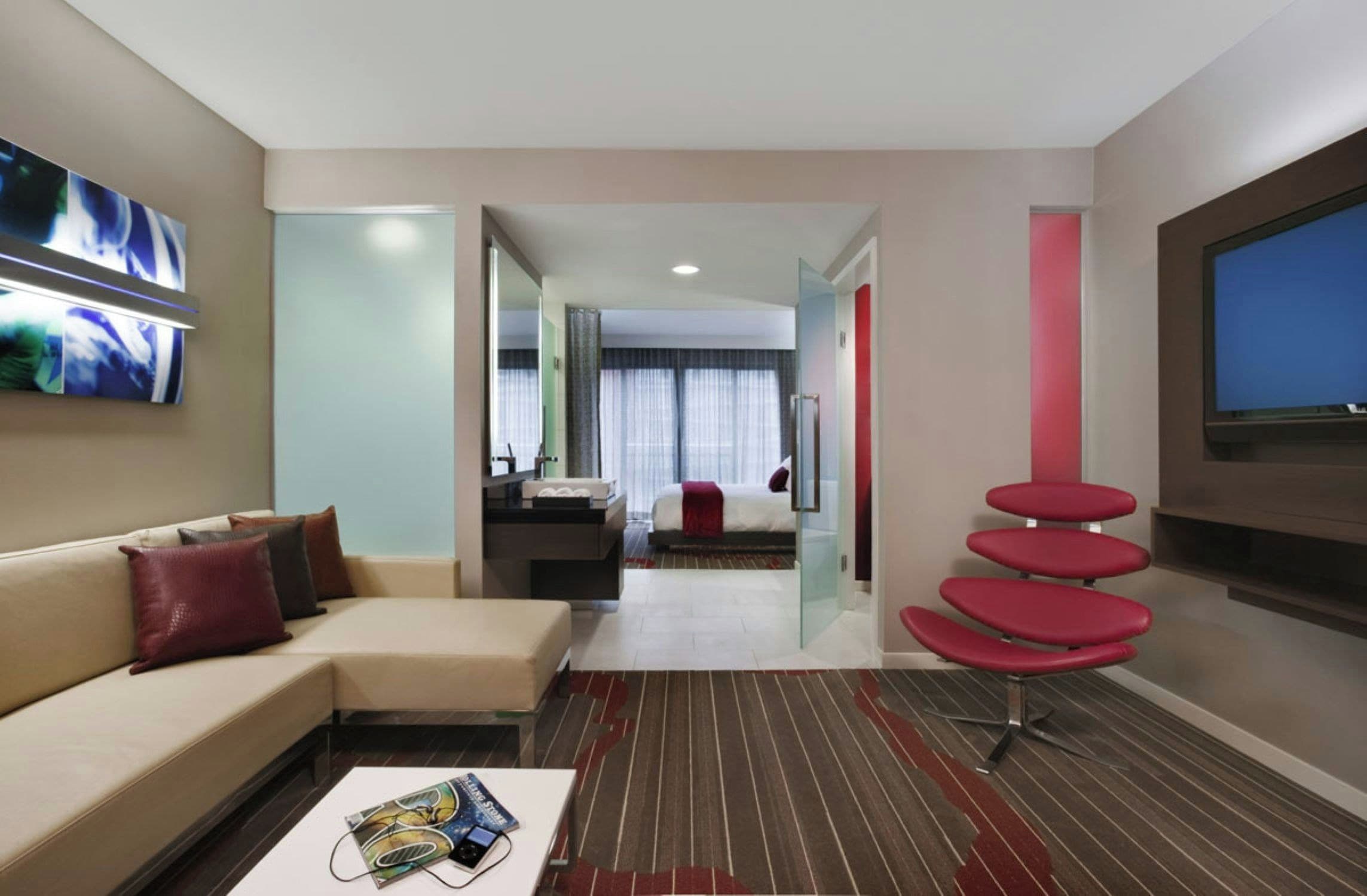 A hotel room with a sofa, a coffee table, a TV, a glass partition, and a red chair.