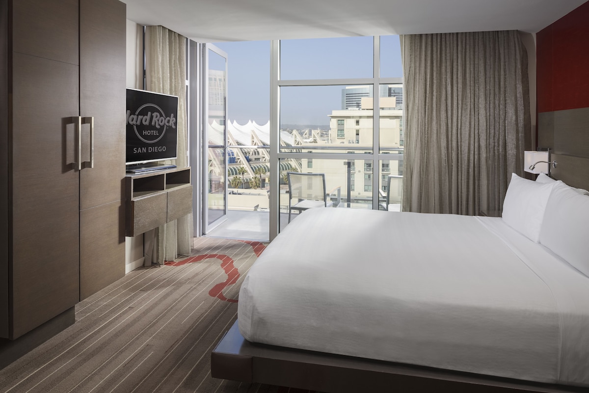 Een hotelkamer met een groot bed, een tv op een standaard en een uitzicht op de stad door grote ramen.
