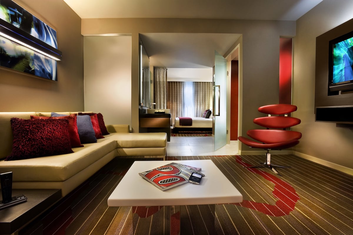 Una habitación de hotel moderna con sofá, mesa de centro, televisor y una zona de estar con una silla roja.