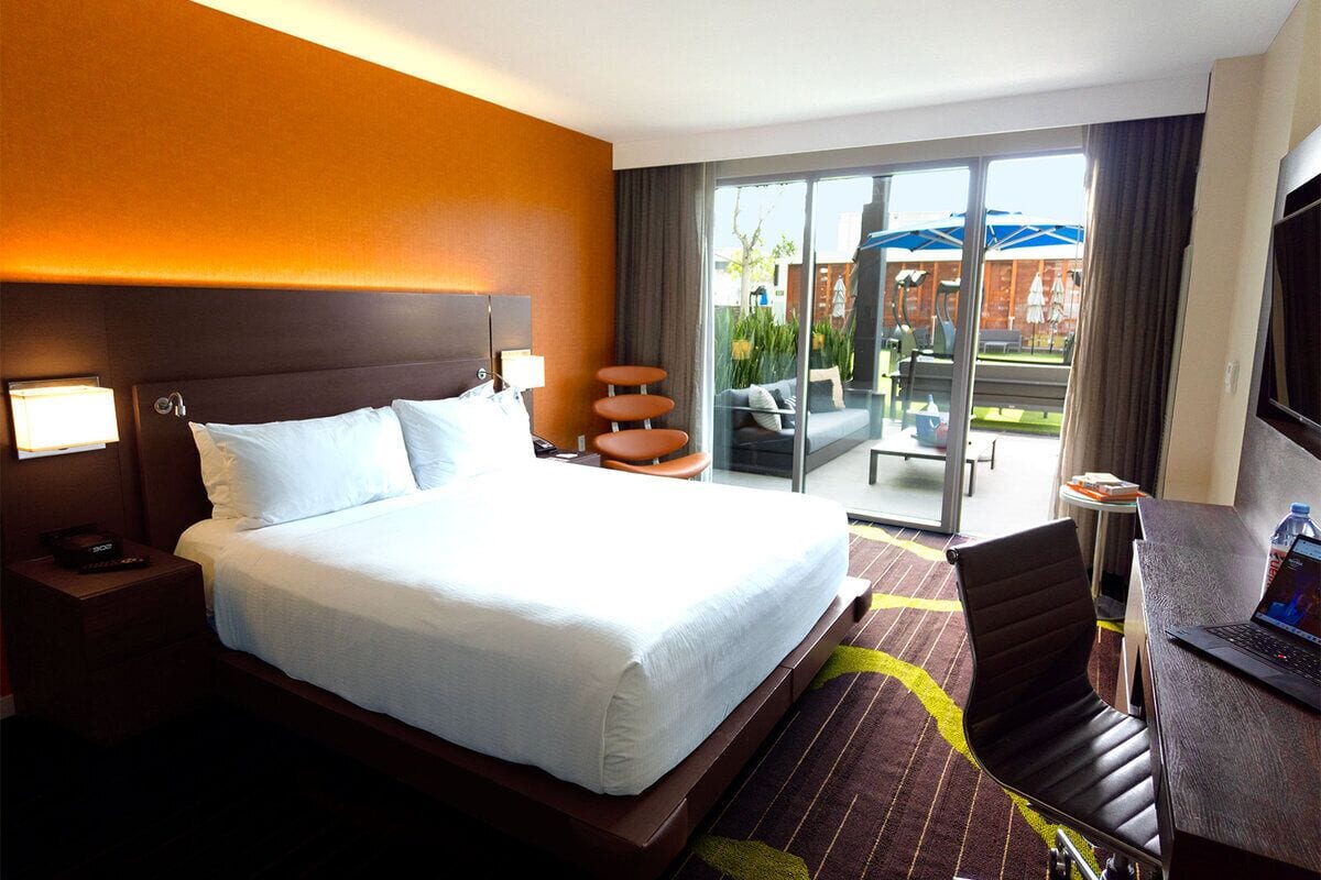 Habitación de hotel con una cama grande, un escritorio con una computadora portátil, un televisor y un balcón con mobiliario exterior.