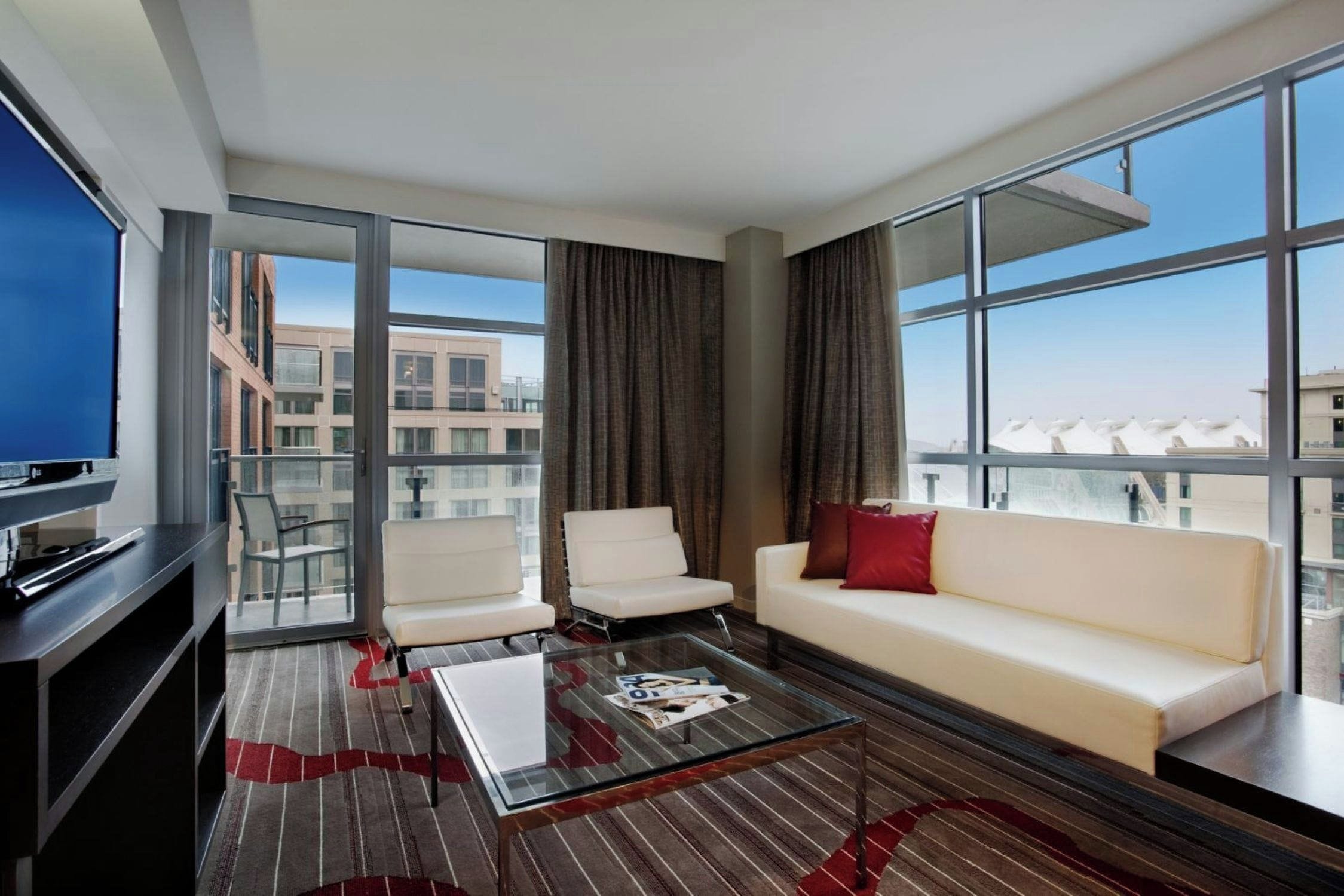 A modern hotel room with a glass partition, a sofa, two armchairs, a coffee table, and a television.