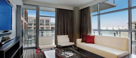 A modern hotel room with a glass partition, a sofa, two armchairs, a coffee table, and a television.