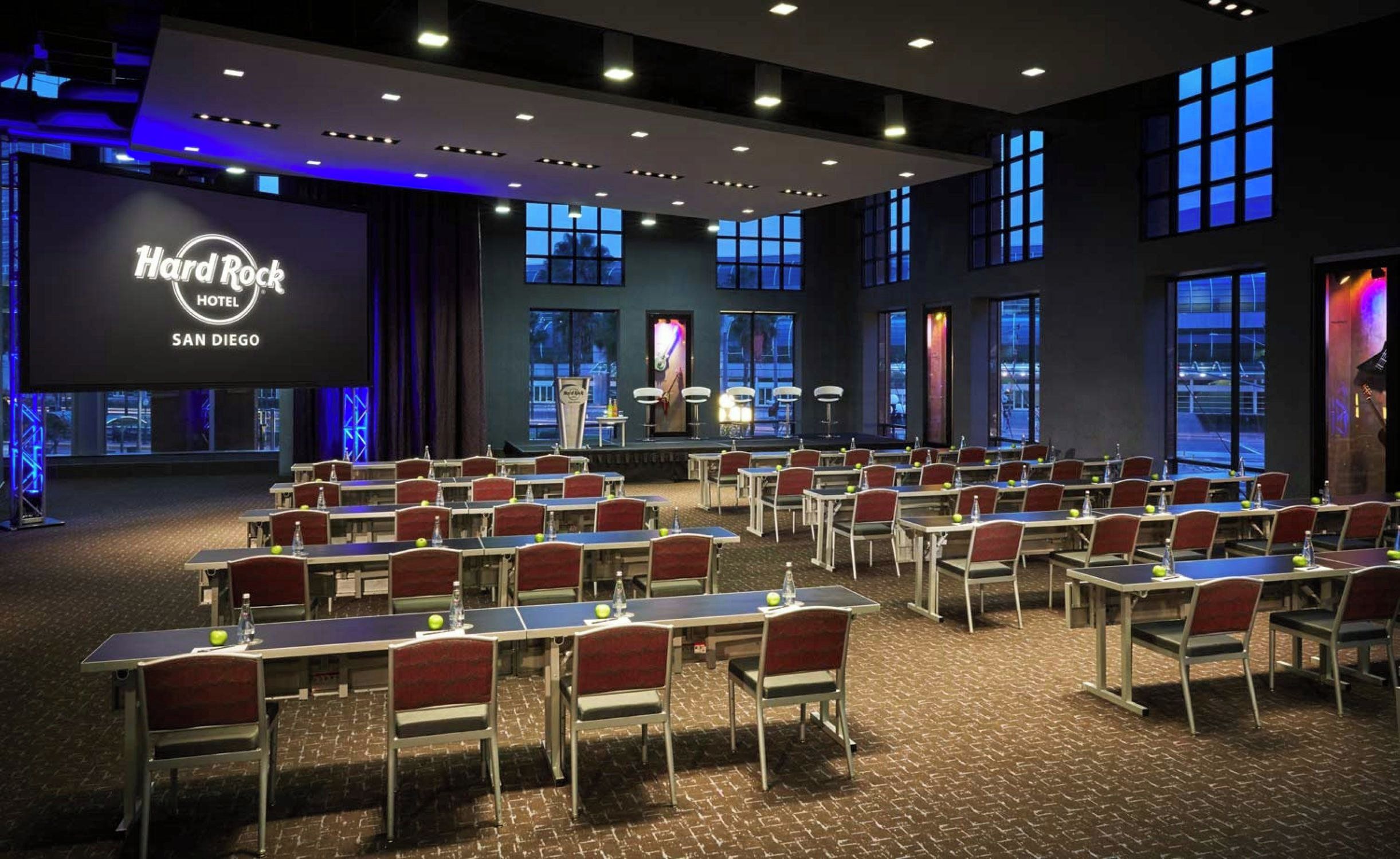 A conference room with tables and chairs, a stage with a presentation screen, and large windows.
