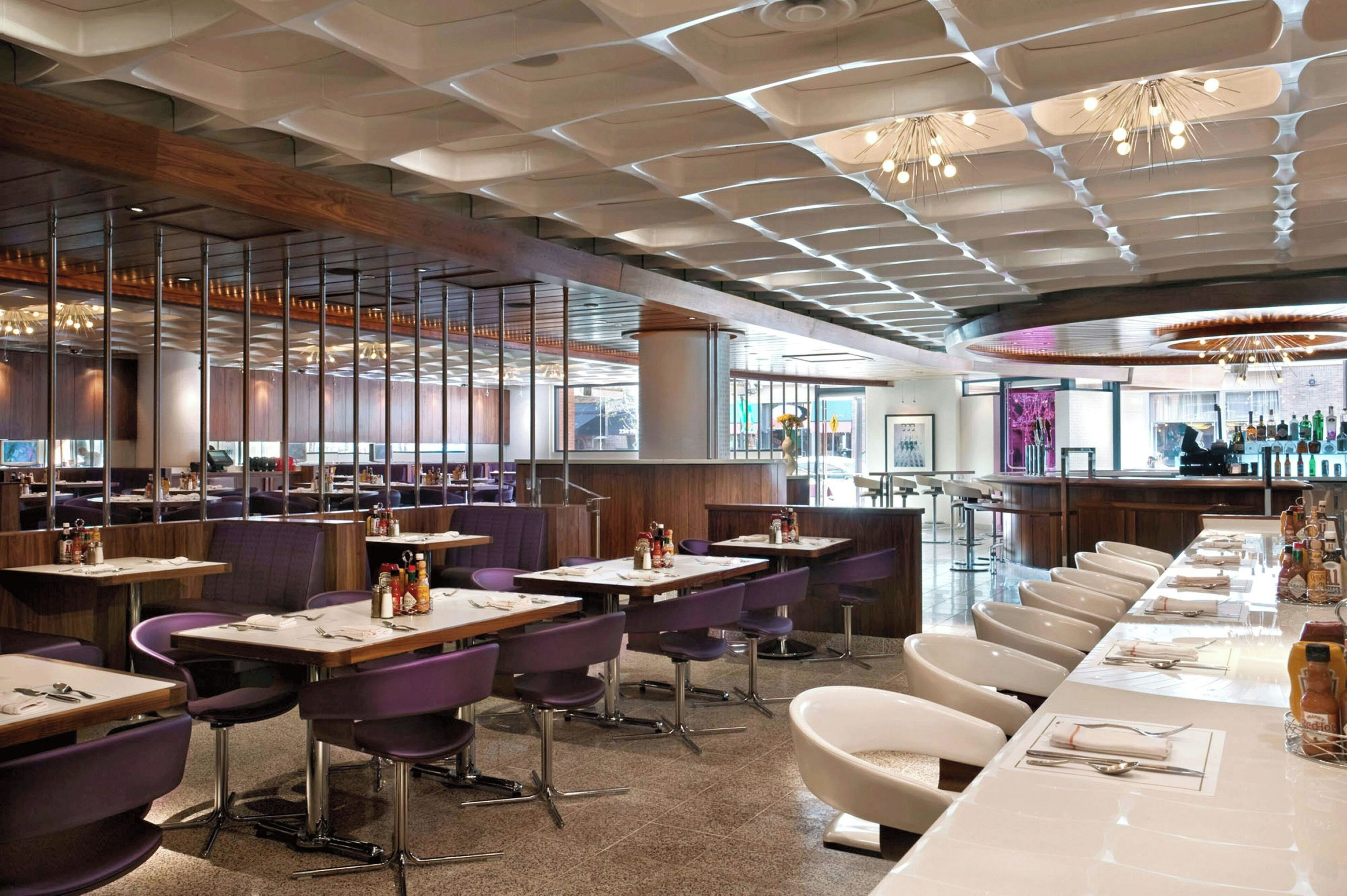 A modern restaurant interior with high ceilings, a long bar, and purple and white seating.