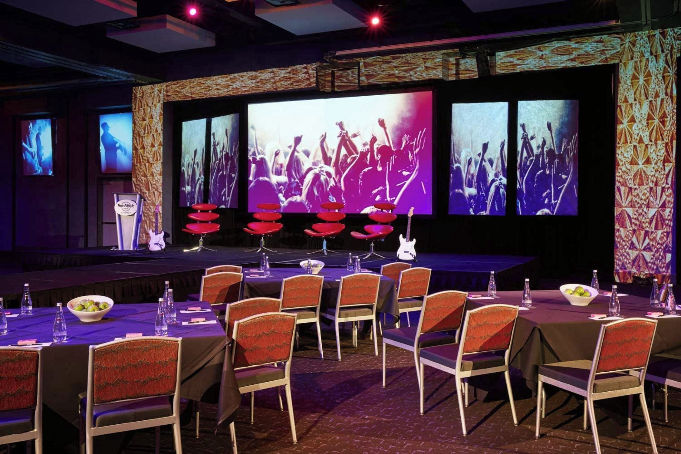 Un salón de conferencias con un escenario, sillas, mesas y una pantalla grande que muestra un concierto.