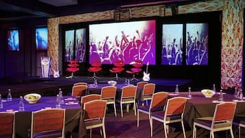 Un salón de conferencias con un escenario, sillas, mesas y una pantalla grande que muestra un concierto.