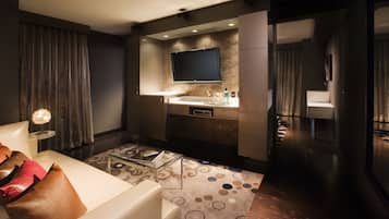 A modern hotel room with a sofa, a glass coffee table, a flat-screen TV mounted on the wall, a small kitchenette, and a patterned rug.