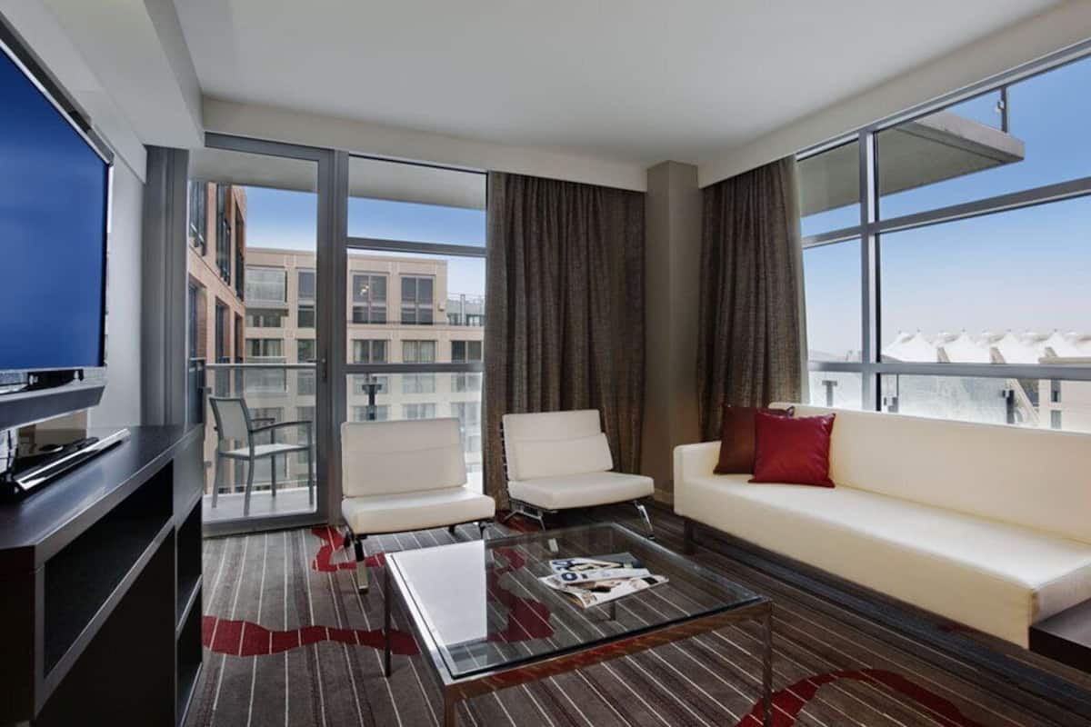 Una habitación de hotel moderna con un ventanal grande, un sofá, dos sillones, una mesa de centro y un televisor sobre un mueble bajo.
