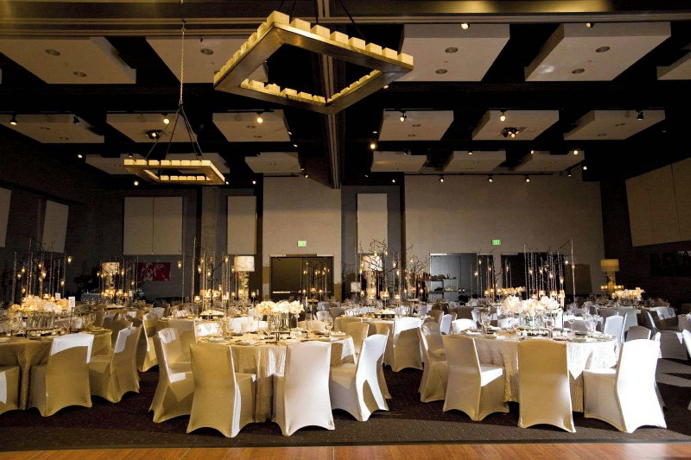 A banquet hall with multiple round tables, white chairs, and overhead lighting fixtures.
