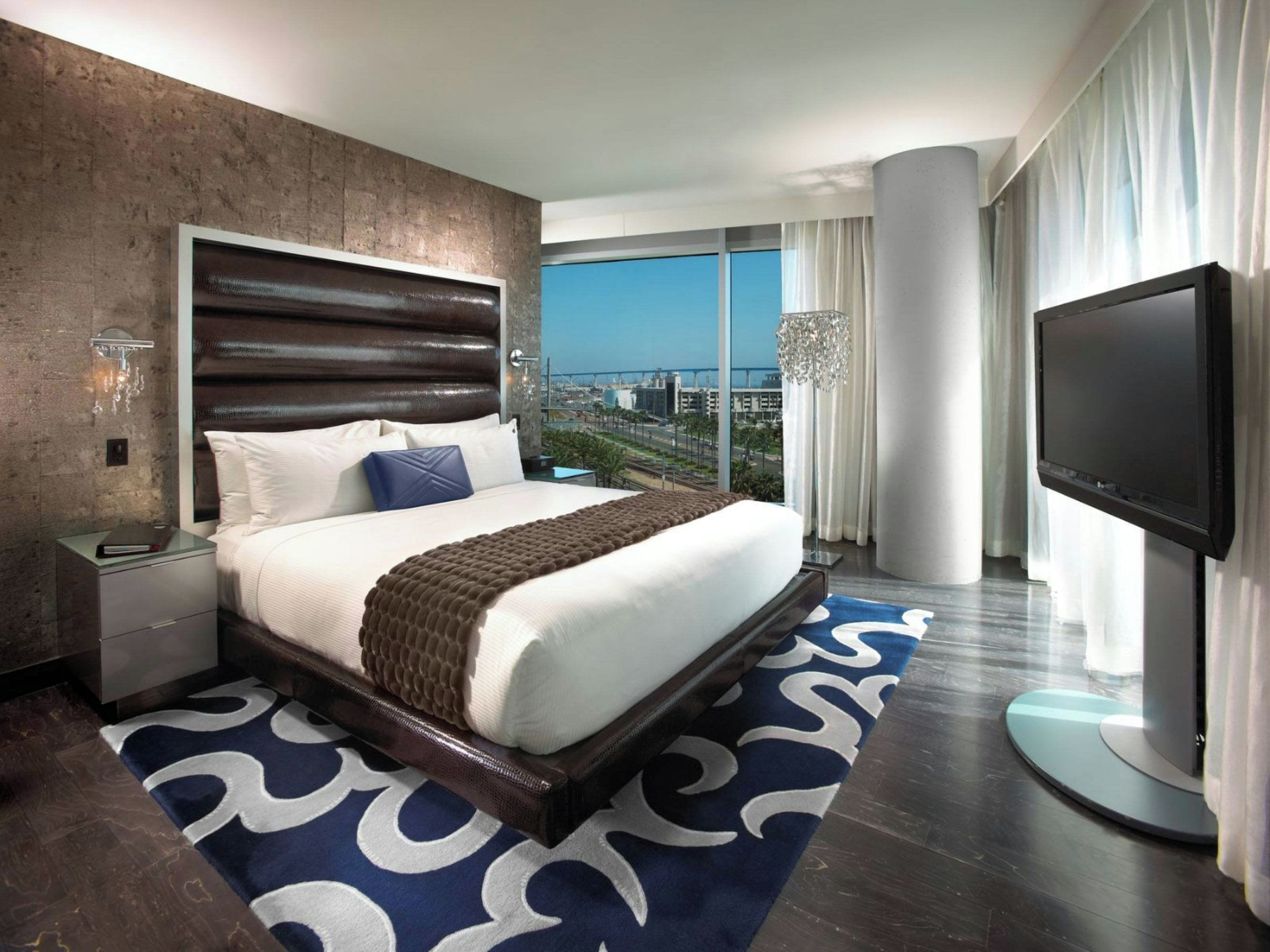 A modern hotel room with a large bed, a television, and a view of the cityscape.