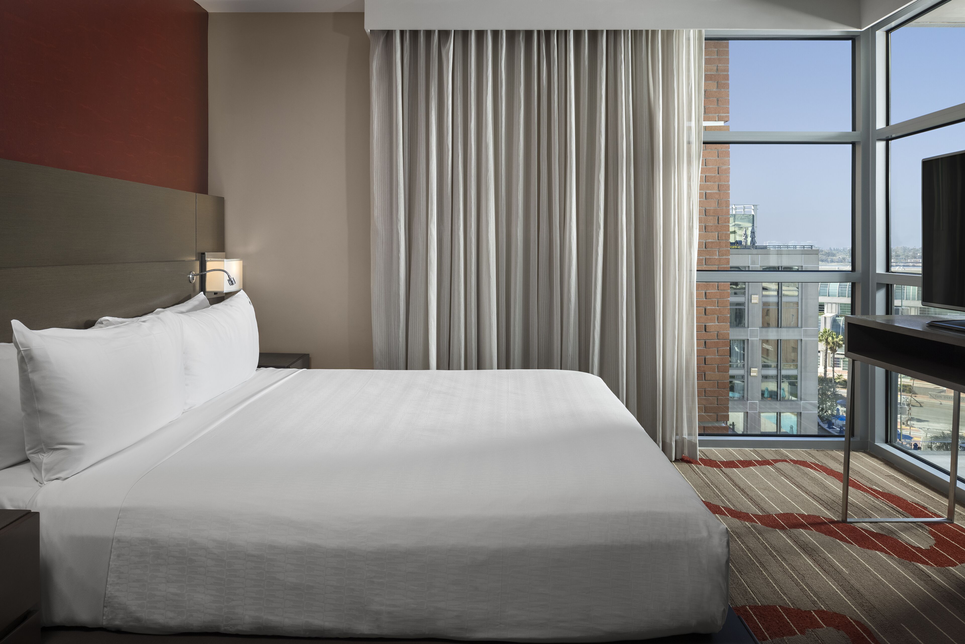 A hotel room with a large bed, a nightstand, a desk with a lamp, a view of the city through large windows, and a striped carpet.