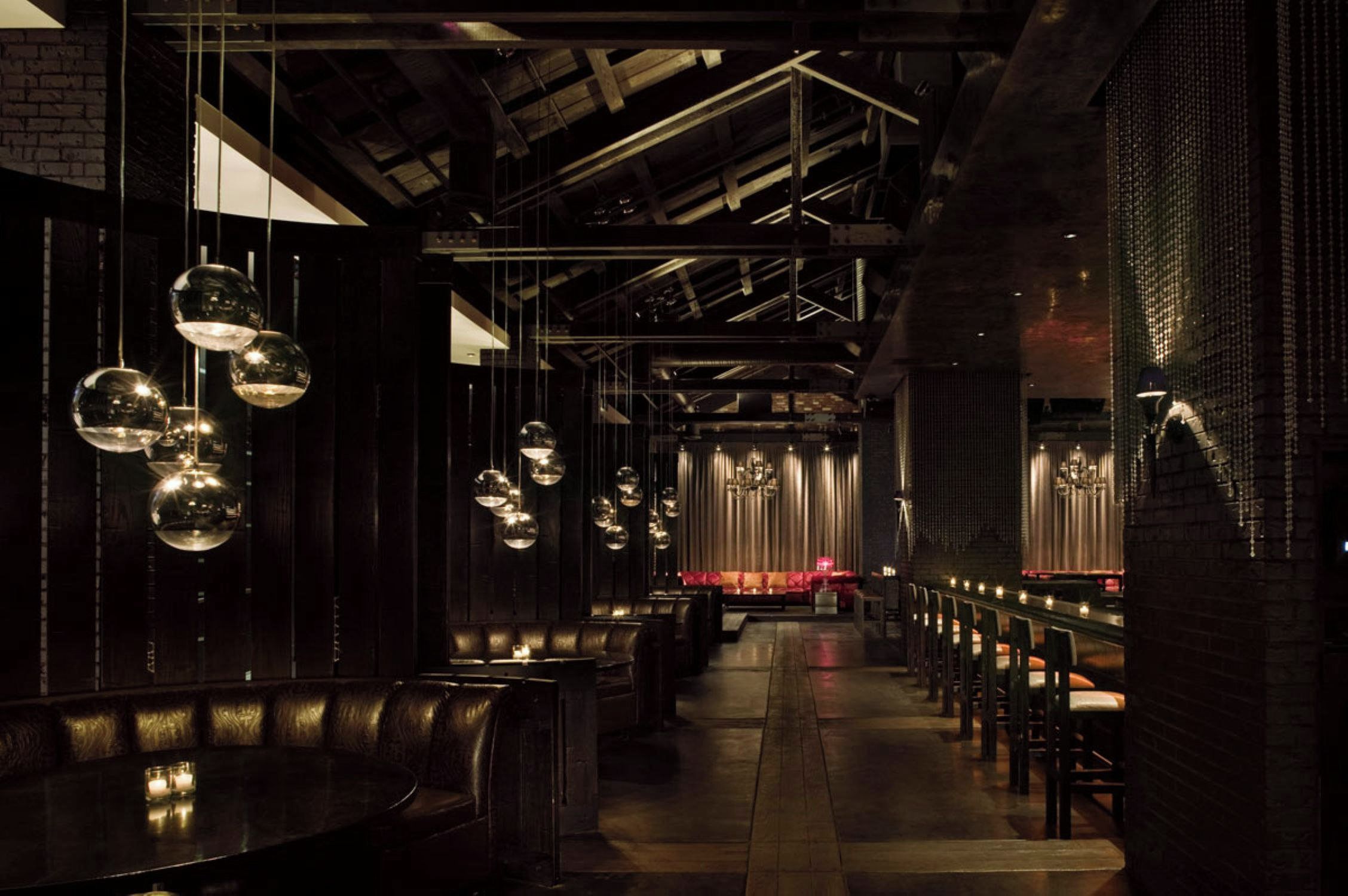 A dimly lit interior with a long bench seating area, pendant lighting, and a stage with curtains.