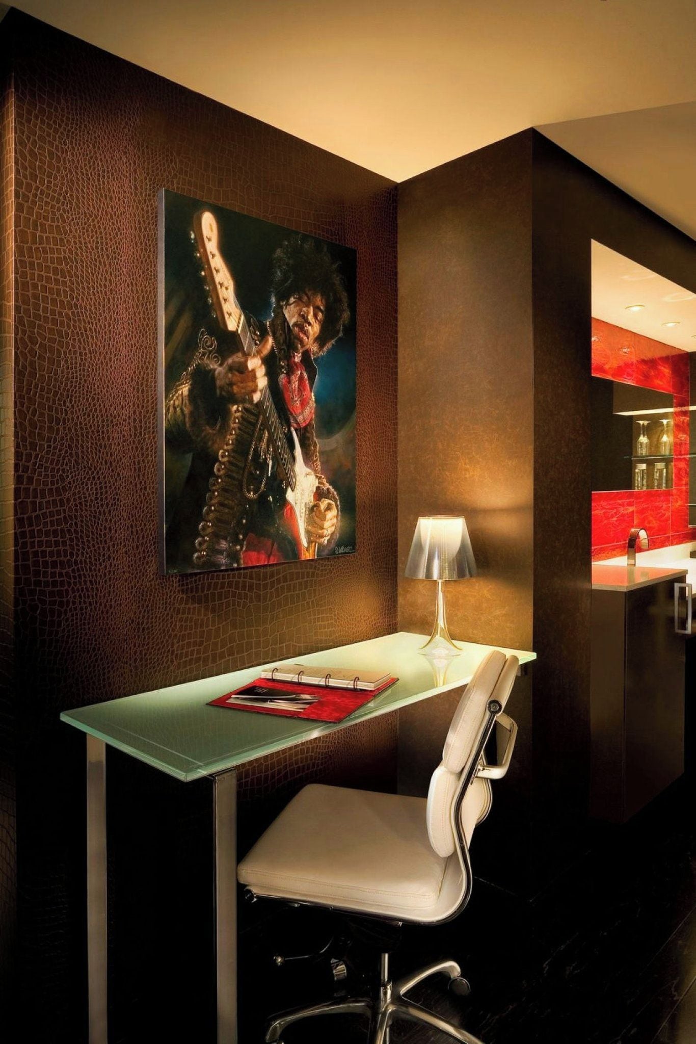 Habitación de hotel moderna con una mesa de vidrio, una silla blanca acolchada, una lámpara y una obra de arte enmarcada en la pared.